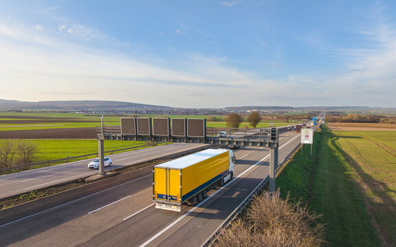 A yellow semi-truck driving on the autobahn under the digital speed limit signs in rural landscape with the blue sky