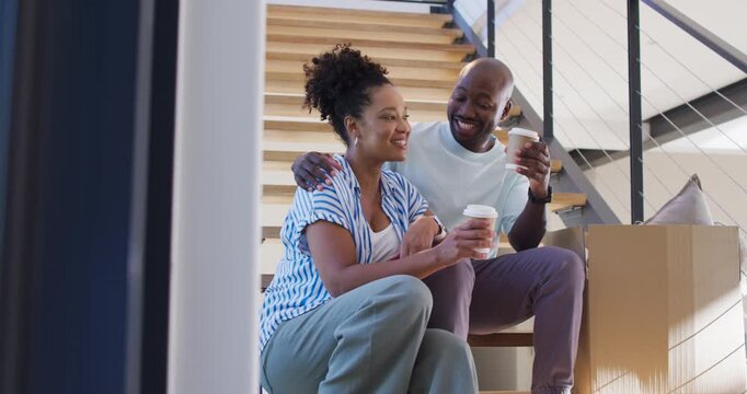 African American couple pausing during move, sitting on lower wood stairs, clinking takeaway cups
