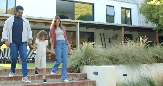Family leaving modern two-story house, holding hands and guiding child down brick steps toward lawn