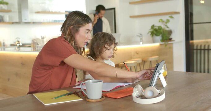 Adult woman leaning, pointing and tapping tablet, guiding child for schoolwork at home dining table