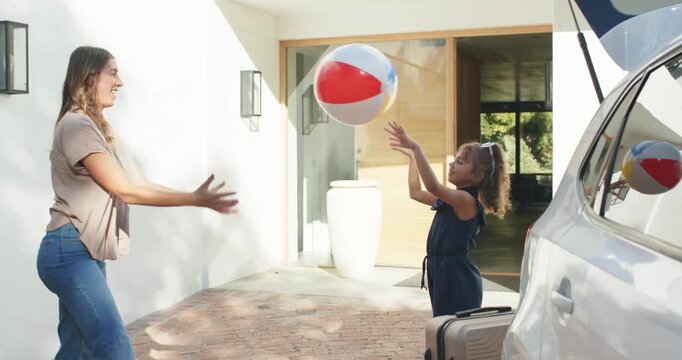 Diverse adult woman and girl tossing large beach ball, girl initiating play before trunk loading