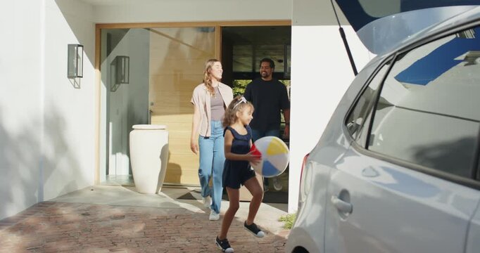 Diverse family child running from doorway on driveway bouncing ball while mom pulling case to car