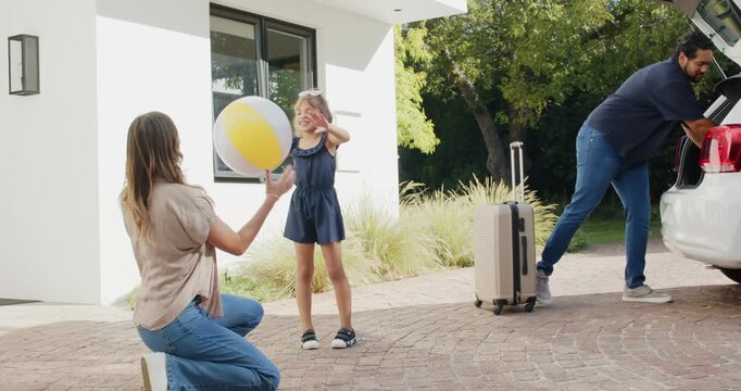 Diverse family packing trip woman kneeling catching beach ball while man loading case on driveway