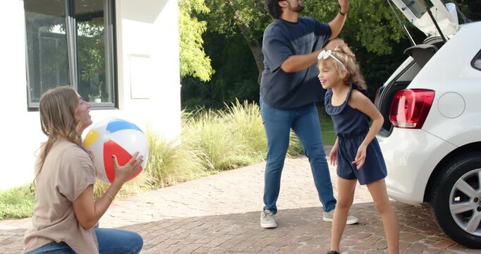 Diverse family passing bright beach ball on driveway by open white hatchback after trunk move