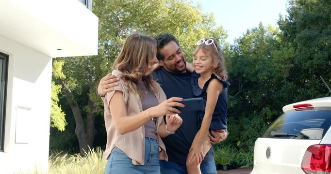 Diverse family huddling on driveway viewing phone, mother holding device, white sunglasses visible