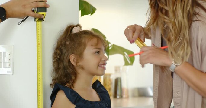Family measuring daughter in kitchen with yellow tape measure, pencil marking head, tracking growth