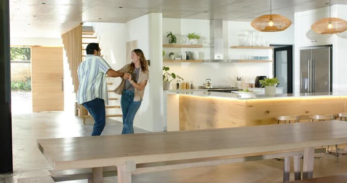 Diverse couple holding hands in kitchen, man reaching her face, leading playful spins to island