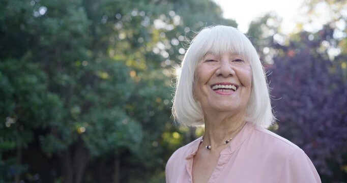 Senior woman in pink blouse, necklace in garden turning, smiling, laughing while sun warming hair