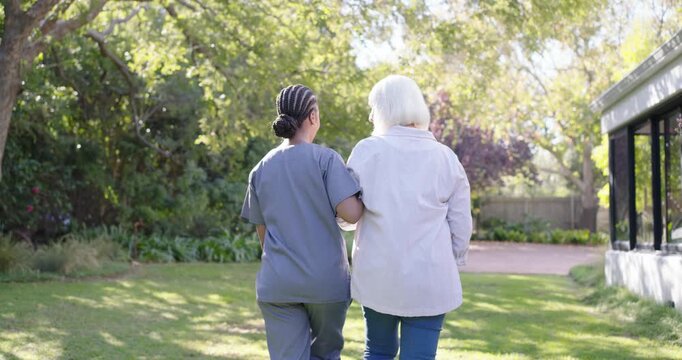Diverse female scrub nurse guiding senior woman arm-in-arm across lawn to veranda, escorting house