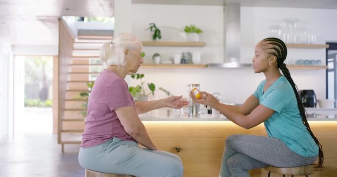 African American carer showing pill bottle, pointing, helping senior woman reading label at kitchen