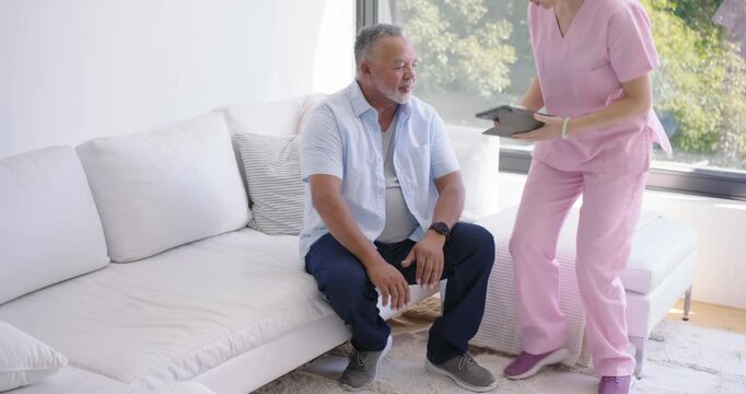 Caregiver woman in pink scrubs reaching for tablet, sitting beside man on sofa, reviewing care plan