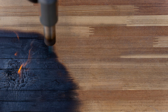 Gas torch burning the edge of a wooden surface, creating a black charred area with visible grain and small flames outdoors.