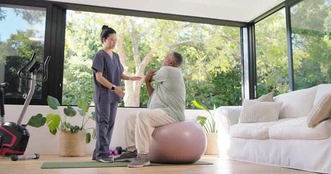 Asian PT in scrubs reaching and guiding senior man on exercise ball arm-cross trunk-turn for rehab