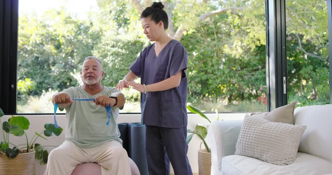 Asian therapist in navy scrubs adjusting grip guiding man pulling band for shoulder rehab at home
