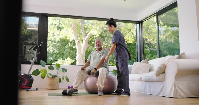 Senior African American man on stability ball curling weight after Asian PT in scrubs guiding rehab