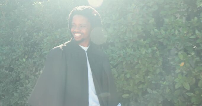 African American man by hedge reacting to off-camera sound, turning, smiling, walking in black robe