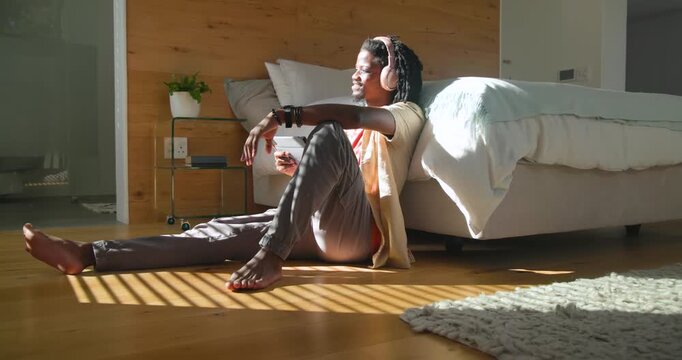 African man with pink headset sitting in bedroom beside bed sunlight moving turning checking phone