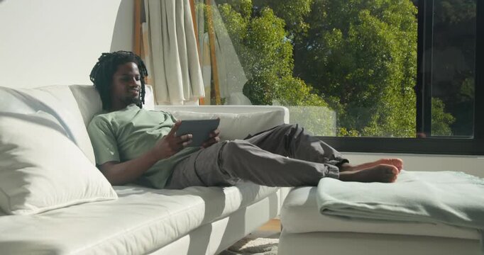 African American man starting with tablet while relaxing on sofa, tapping and glancing out window