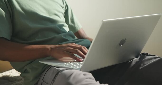 Closeup framing African American man typing on silver laptop in armchair by window pausing thinking