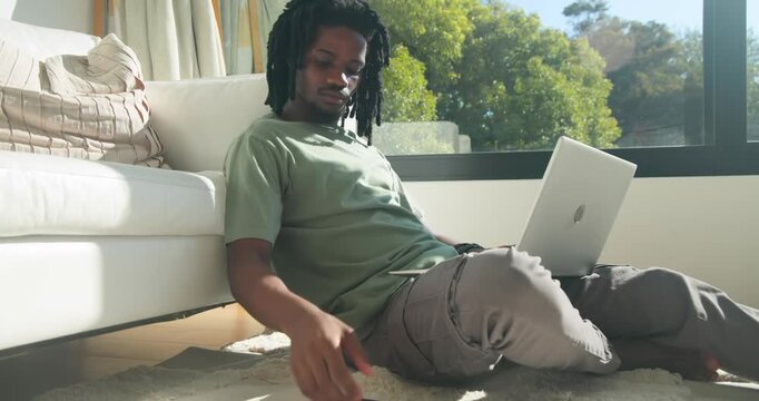 African man sitting on rug by sofa in green tee, typing silver laptop, answering incoming phone