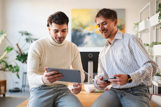 Two men are sitting on a desk, one holding a tablet and the other a smartphone, engaged in a discussion in a modern office setting.