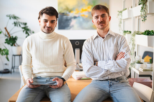 Two young men sit on a desk in a modern office, one holding a tablet and the other a smartphone, engaged in their work.