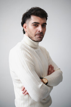 A man with dark hair and a beard wears a white turtleneck sweater and a watch. He stands with his arms crossed against a plain background.
