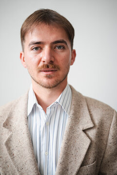 A young man with light brown hair and a mustache wears a striped shirt and a tweed jacket, looking directly at the camera.