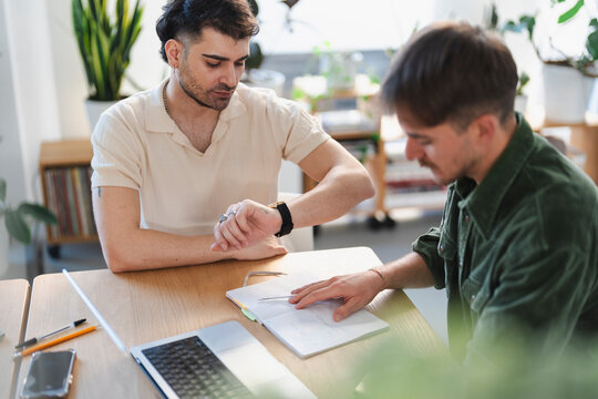 Two men collaborate at a desk, one checking his watch while the other points to notes in a notebook, surrounded by office supplies and a laptop.