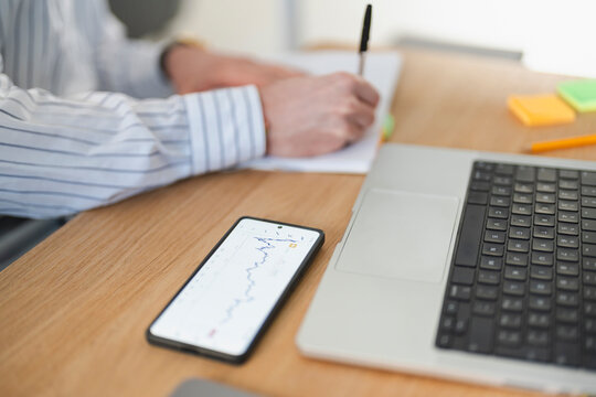 A person in a striped shirt writes in a notebook while a smartphone displays a stock chart and a laptop sits nearby on a wooden desk.