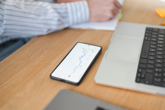 A person is working at a desk, reviewing financial data on a smartphone and taking notes in a notebook.