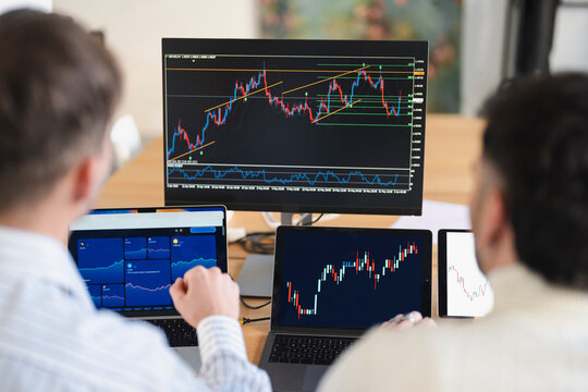 Two individuals analyze financial market data displayed on multiple screens, focusing on trading charts and stock performance.