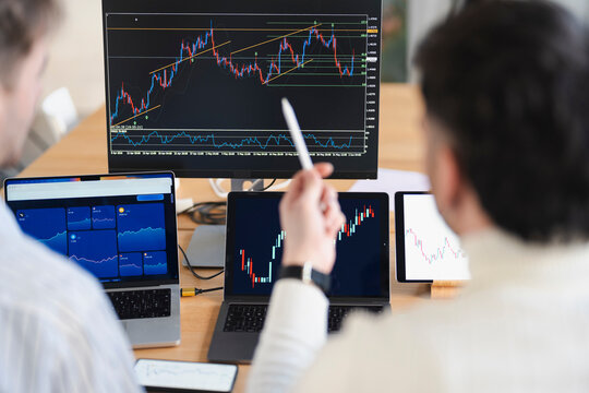 Two individuals analyze financial charts on multiple screens, discussing market trends and investment strategies.