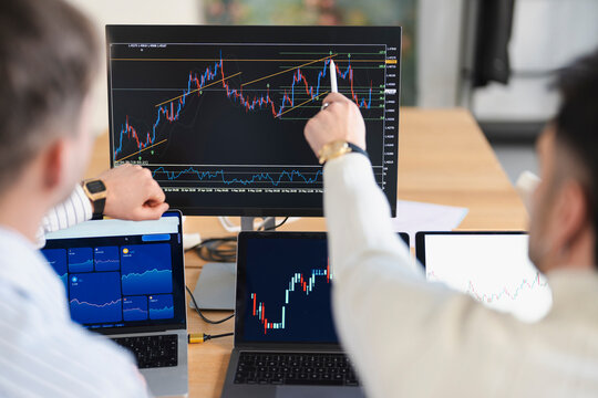 Two traders analyze financial charts on multiple screens, one pointing to a specific point on a large monitor, indicating a discussion about market trends and investment strategies.