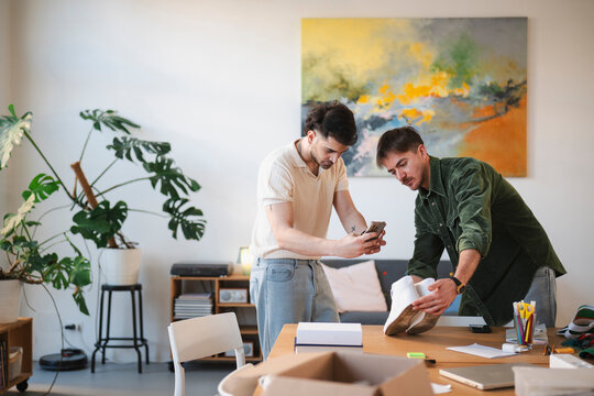 Two young men are in a bright room, one holding a phone and the other holding a pair of white shoes, examining them closely.