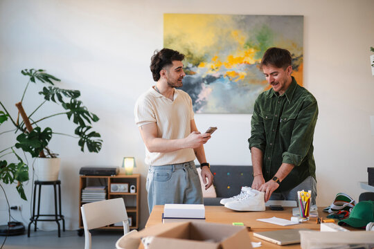 Two men are in a bright room, one holding a phone and the other examining a pair of white sneakers on a wooden table.