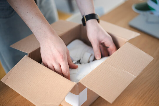A person's hands carefully place a new pair of white sneakers into a cardboard shipping box on a wooden table.