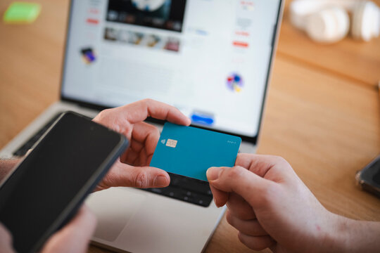 A person holds a debit card and a smartphone while a laptop displays a website, suggesting online banking or shopping.