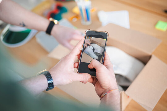 A person photographs new white sneakers inside a cardboard box with their smartphone, capturing the moment of unboxing a recent purchase.