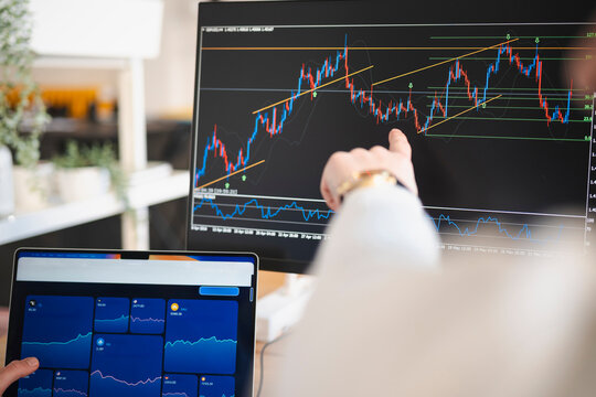 A person points at a financial chart on a computer screen, analyzing market data. Another screen displays various investment options and their performance metrics.