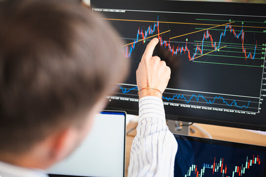 A person analyzes financial charts on multiple computer screens, pointing to a specific point on a candlestick graph.