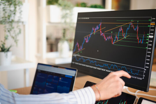 A person analyzes financial charts on multiple screens, pointing at a stock market graph on a large monitor.