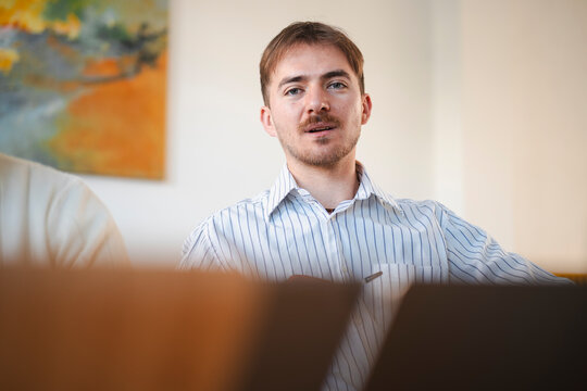 A young man in a striped shirt sits at a table, looking forward with an open mouth. He is in an office setting with a painting on the wall.