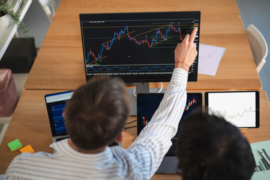 Two individuals analyze financial charts on multiple screens, one person pointing to a complex trading graph on a large monitor.
