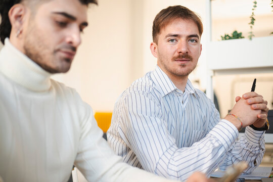 Two men are in a meeting, one looking directly at the camera while the other is out of focus. They are seated at a table with papers and a pen.