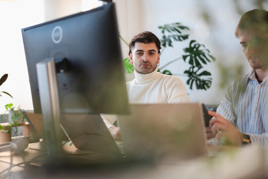 Two men work together in a bright office setting, one looking directly at the camera while the other is focused on a laptop.