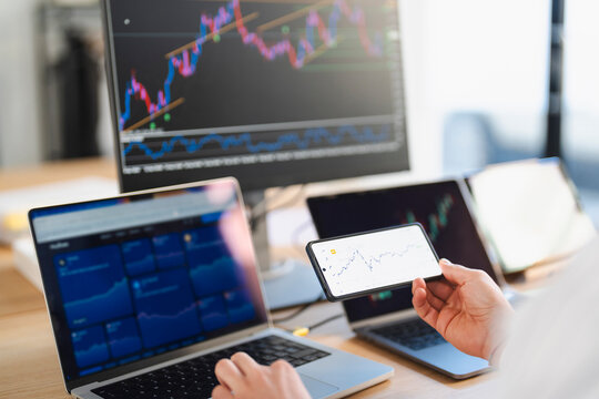A person analyzes financial charts on multiple screens, including a laptop, a large monitor, and a smartphone, indicating active trading or investment research.