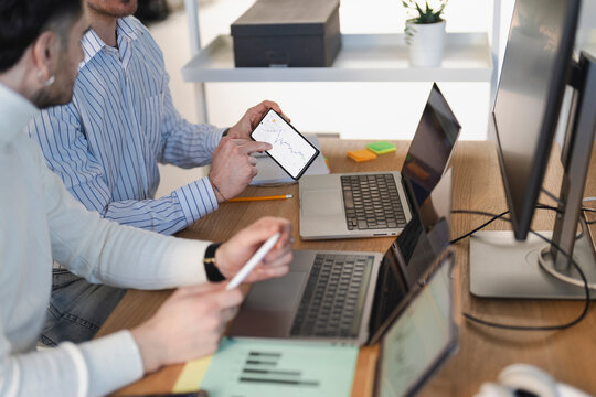 Two men collaborate at a desk, analyzing financial data on a smartphone and laptops, discussing business strategy in a modern office environment.