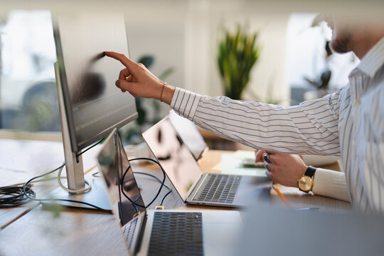 A man in a striped shirt points at a computer screen while working on multiple laptops at a desk.