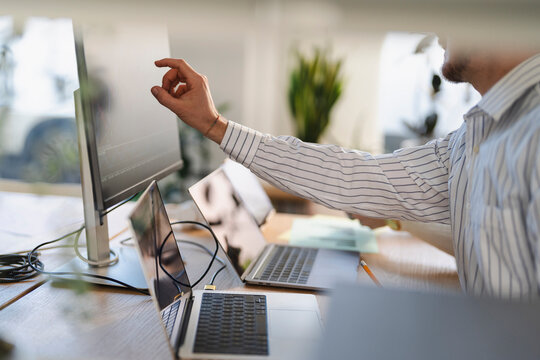 A man in a striped shirt points at a computer screen while working at a desk with multiple laptops and a plant in the background.